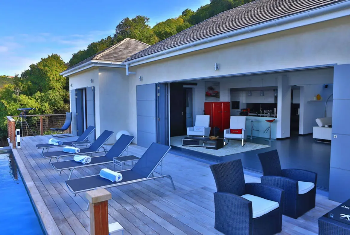 Four blue lounge chairs with rolled white towels line the wooden deck beside a crystal pool, while dark wicker armchairs and a telescope invite endless sky-gazing. The villa's beige facade with blue doors opens wide to reveal the red fridge and modern living area within—indoor-outdoor perfection.