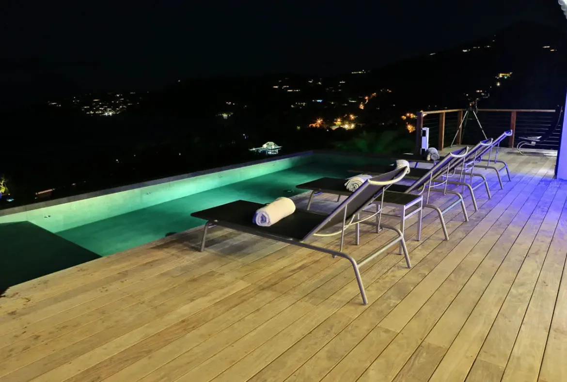 Five lounge chairs with rolled white towels line the wooden deck beside a pool glowing green under underwater lights. Distant city lights twinkle across the dark hills while blue accent lighting traces the terrace—night swimming with a view of St. Barths at your feet.