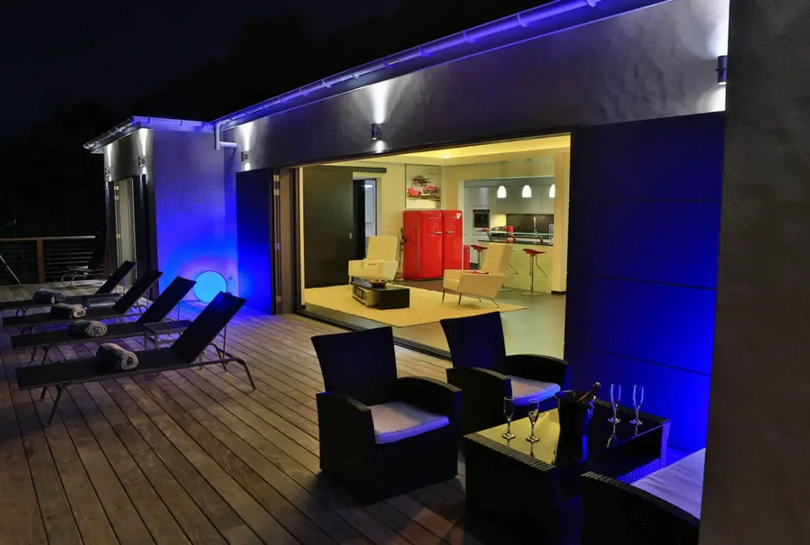Black lounge chairs with rolled white towels line the wooden deck, while two wicker armchairs surround a side table set with champagne flutes and a chilled bottle. Blue LED strips illuminate the exterior walls as the indoor kitchen glows warmly beyond—Caribbean nights were made for this.