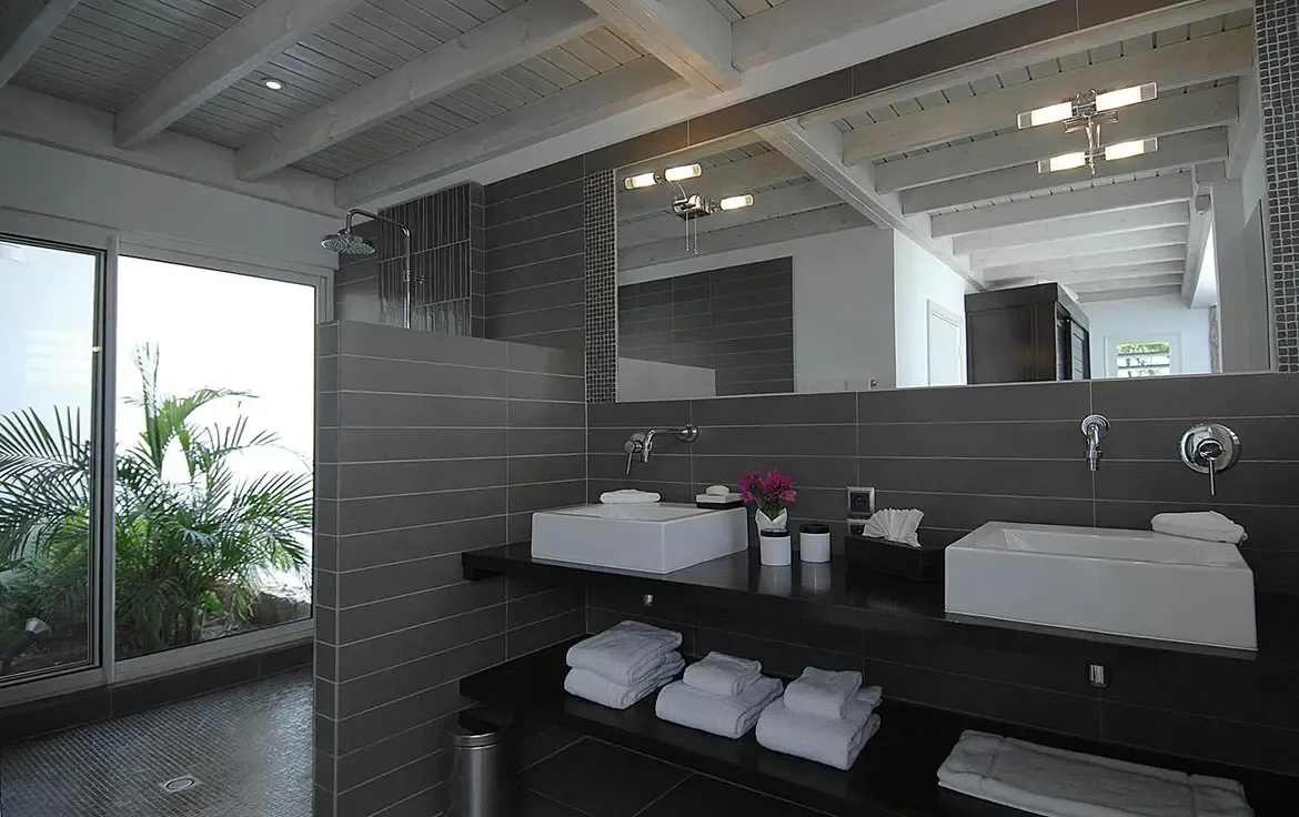 Two white vessel sinks sit on a black floating vanity under a frameless mirror, where a glass door in the shower reveals lush tropical palms and purple flowers add a pop of color.