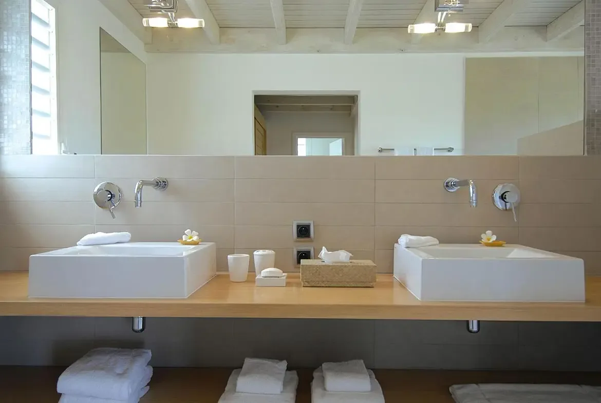 Two white square vessel sinks sit on a light wood floating vanity, where frangipani flowers in yellow dishes and a wall-to-wall mirror brighten the modern bathroom.