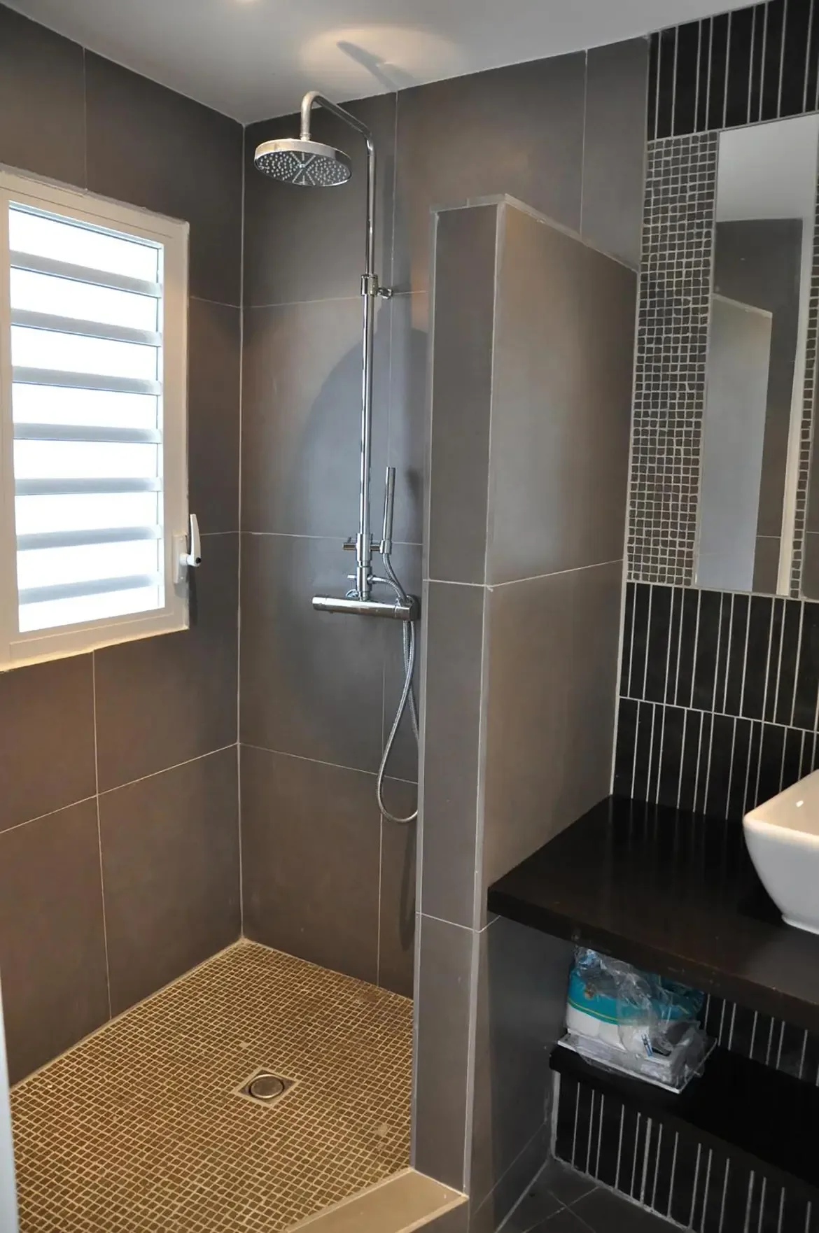 A chrome rainfall showerhead sits behind a grey tile half-wall, where a white vessel sink with pinstripe mosaic tiles and a louvered window add modern elegance.