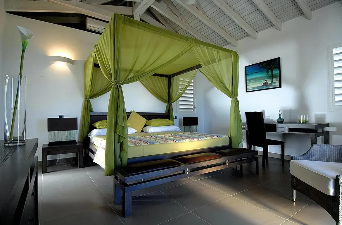 A lime-green canopy bed with sheer drapes sits on grey tiles under a vaulted ceiling, where a calla lily vase and a beach painting with desk add tropical elegance.