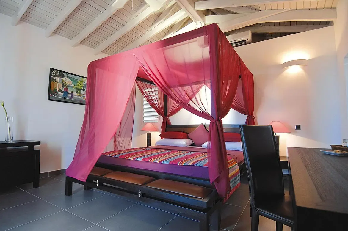 A fuchsia canopy bed with sheer drapes and a multicolored striped runner sits under a vaulted ceiling, where a tropical painting and pink-shaded lamps add bold color.