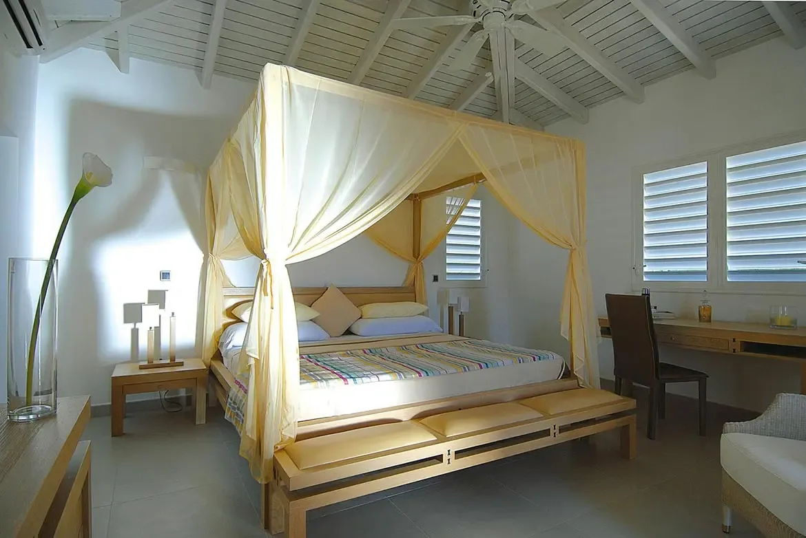 A pale-yellow canopy bed with tied-back sheer drapes sits under a white vaulted ceiling, where a calla lily vase and a wooden bench with cream cushions warm the sunlit room.