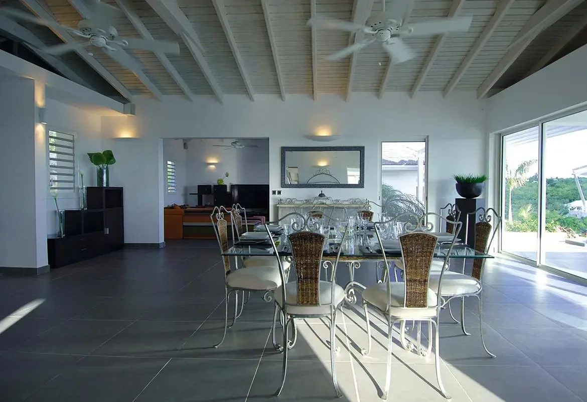 A glass-top dining table surrounded by white wicker chairs sits on charcoal tiles under a vaulted ceiling, where open doors lead to a sunlit terrace and green hillside.