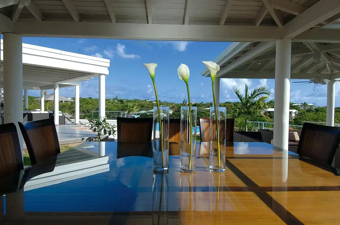 A polished wood dining table with calla lily vases sits under a white pavilion, where dark chairs frame an infinity pool and lush tropical hills beyond.