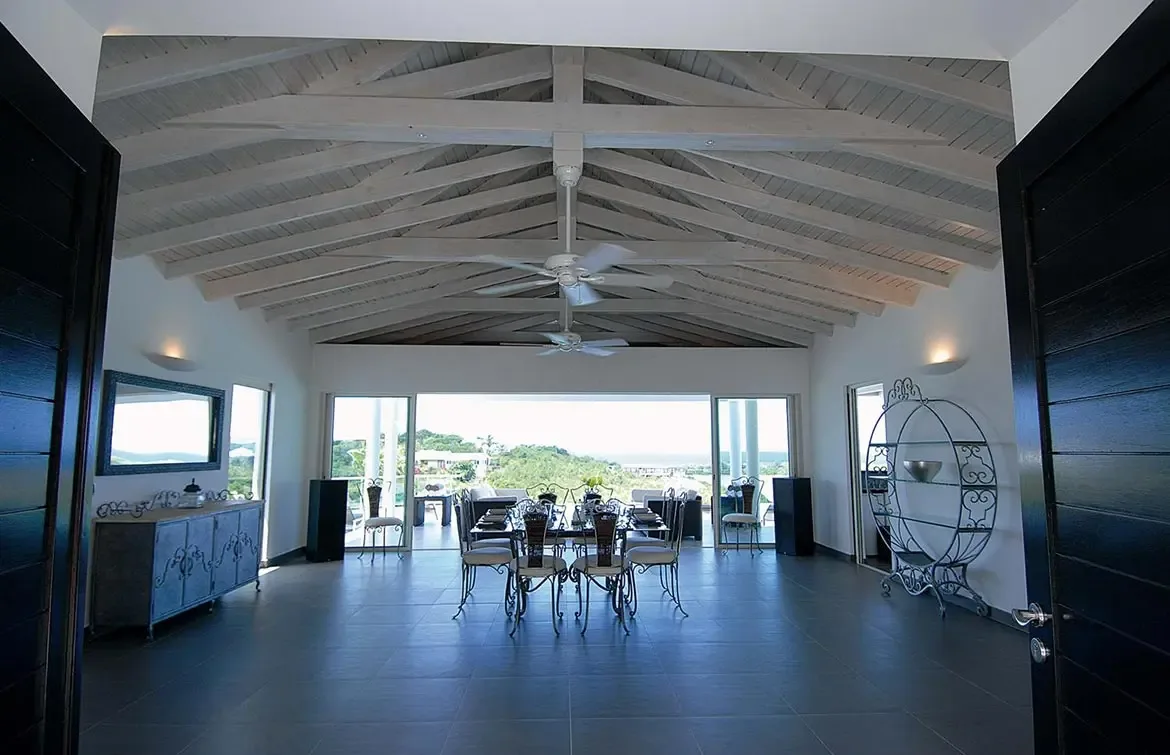 Wrought-iron dining chairs surround a dark table under a vaulted ceiling, where a distressed blue sideboard and large speakers frame open doors to a terrace with ocean views.