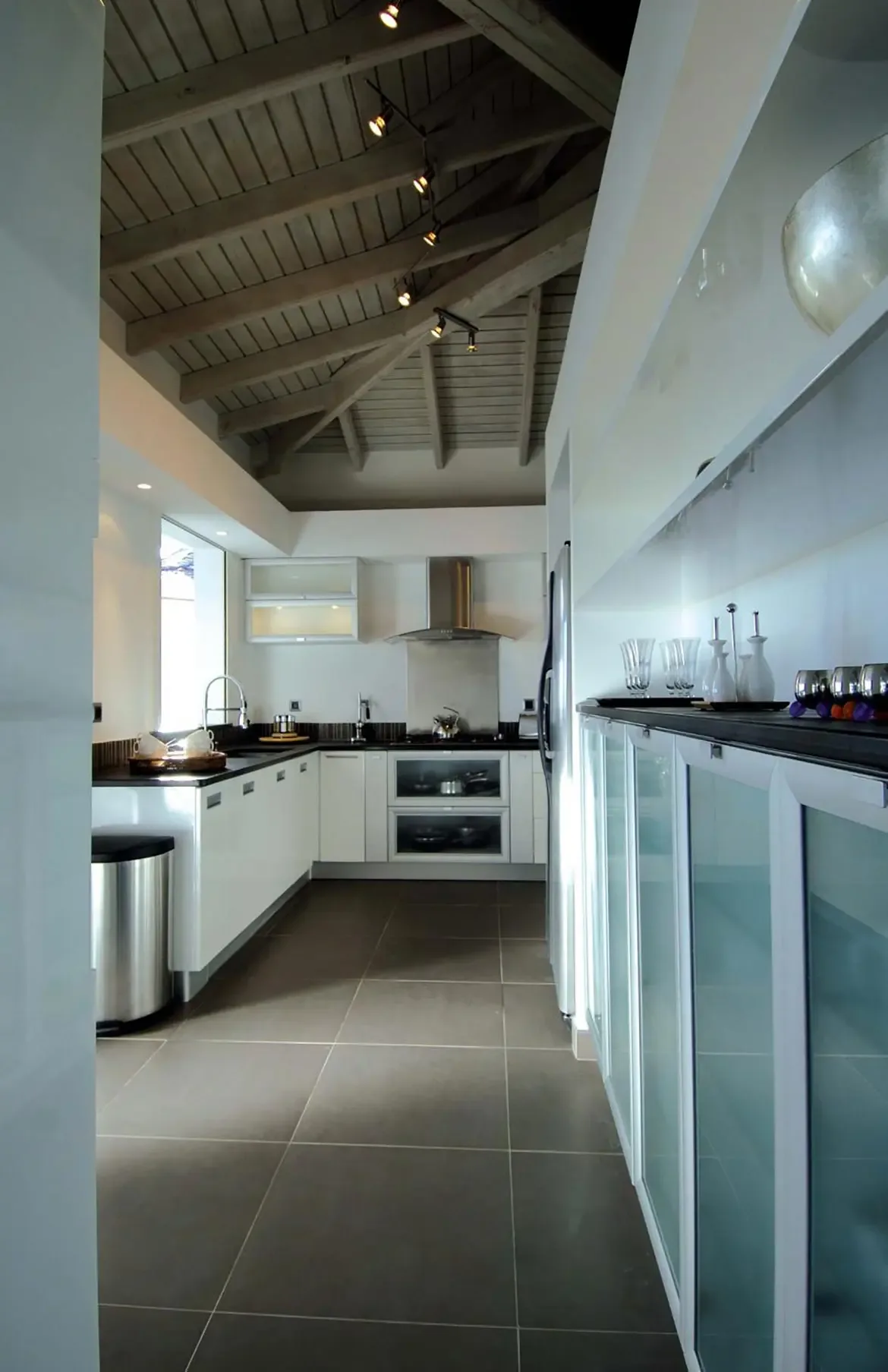 Glossy white cabinets and dark countertops line a galley kitchen under a vaulted wood ceiling, where a double wall oven and stainless hood anchor the modern space.