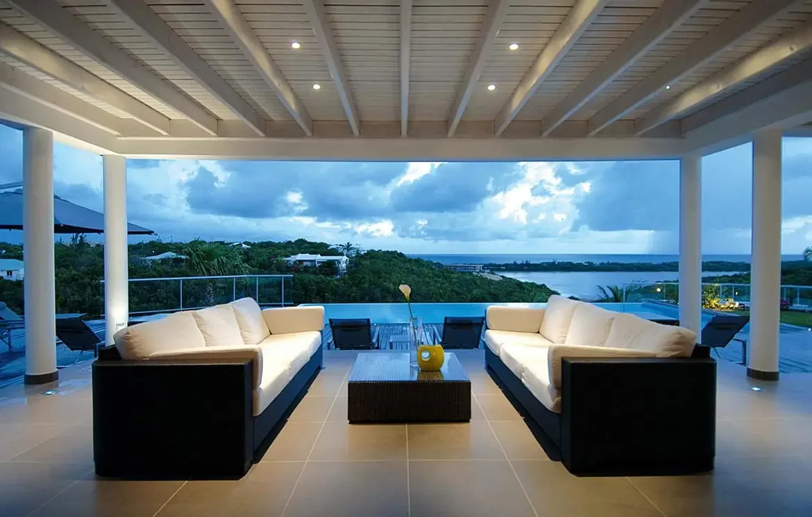 Two dark wicker sofas with white cushions face each other under a white-beamed ceiling, where a glass vase with calla lily sits on a coffee table and dusk falls over the infinity pool.