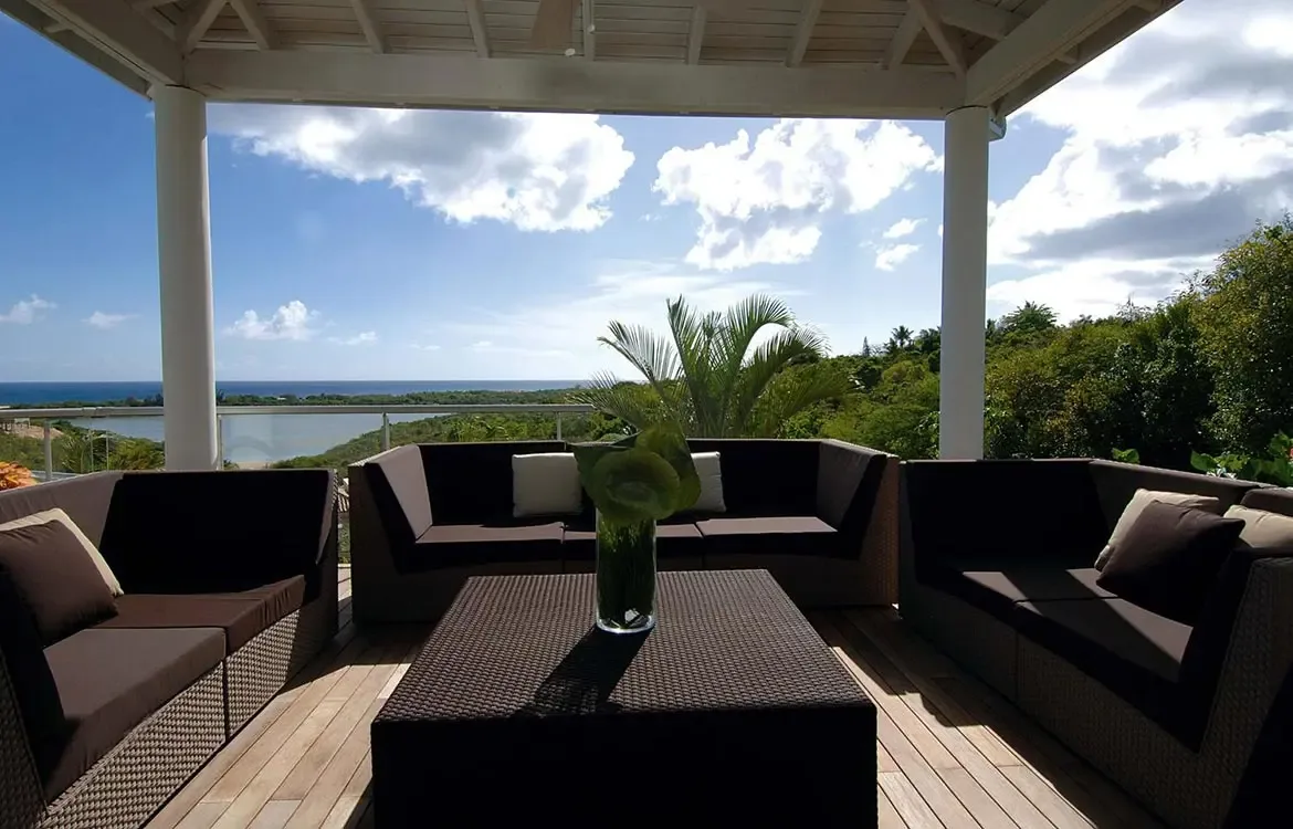 Deep chocolate-cushioned L-shaped sofas sit on a wooden deck under white columns, where a glass vase of green foliage centers the space and a vast ocean panorama unfolds beyond the railing.
