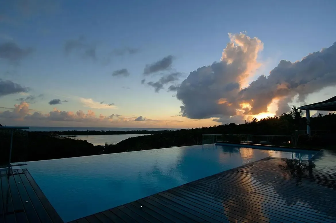 At dusk, floor lights illuminate a wet wooden deck beside a still infinity pool, where sunset clouds glow gold and pink over dark hills and a calm lagoon.