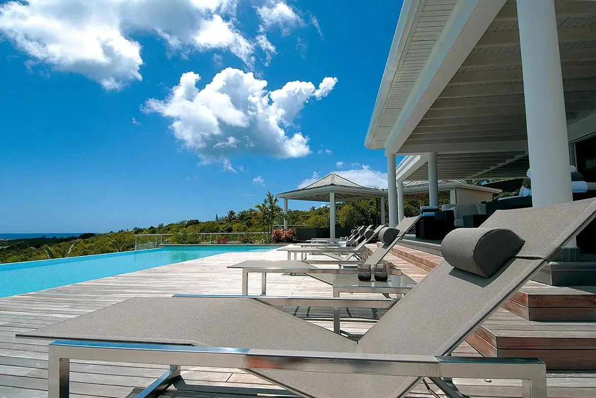 Sleek loungers line a wooden deck beside a turquoise infinity pool, where a white villa with covered verandas and a gazebo overlook lush tropical hills.