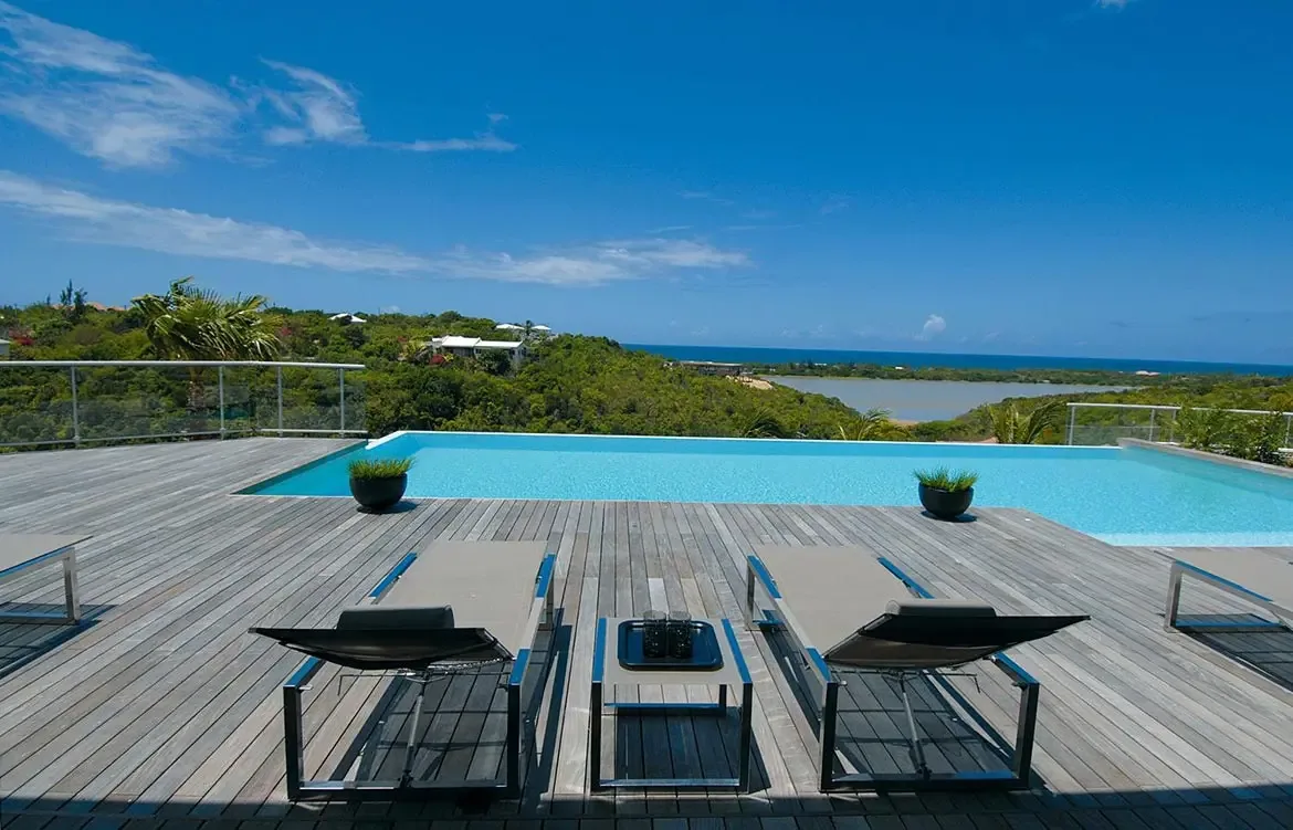 Two grey mesh loungers sit on a weathered deck beside a turquoise infinity pool, where glass railings frame lush hills and a distant lagoon under a blue sky.
