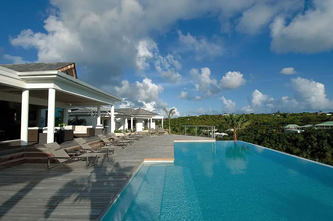 Wide steps descend into a turquoise infinity pool on a weathered deck, where white pillars and a modern villa frame panoramic views of lush green hills and ocean.