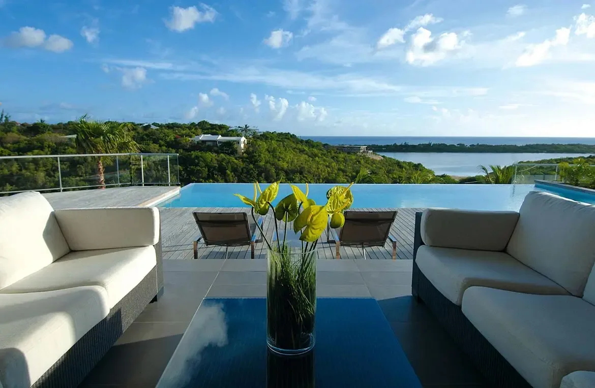 Two white-cushioned sofas face each other across a glass table with yellow anthuriums, where an infinity pool merges with green hills and ocean beyond.