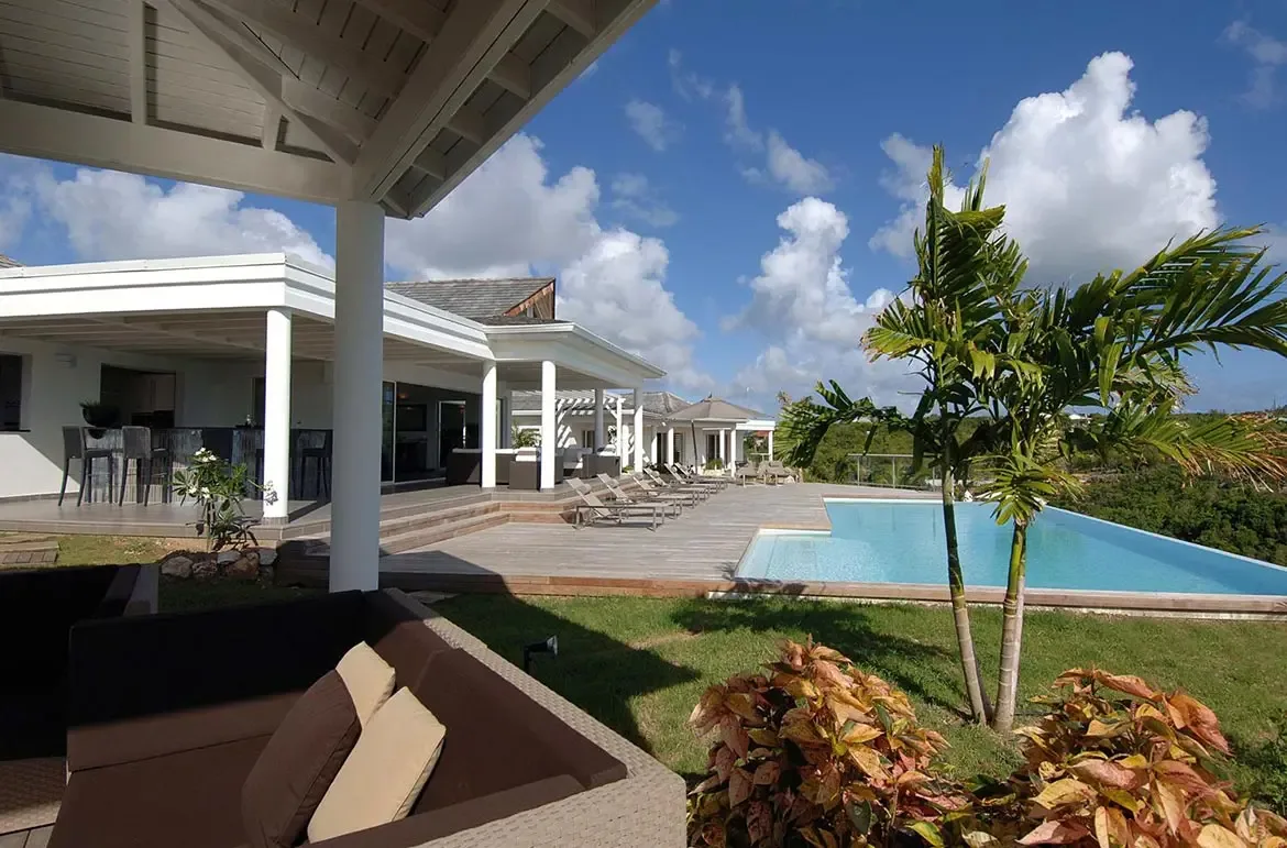 A wicker sofa with white cushions frames a turquoise infinity pool, where a palm and orange tropical shrubs accent the deck above rolling green hills.