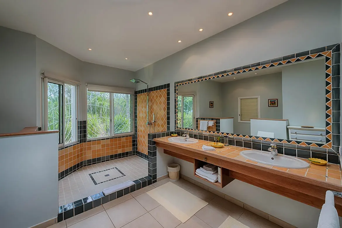 Amber square tiles top a thick wood double vanity beneath a geometric-mirror border, where a walk-in shower with matching amber walls and green-garden windows floods the space with light.