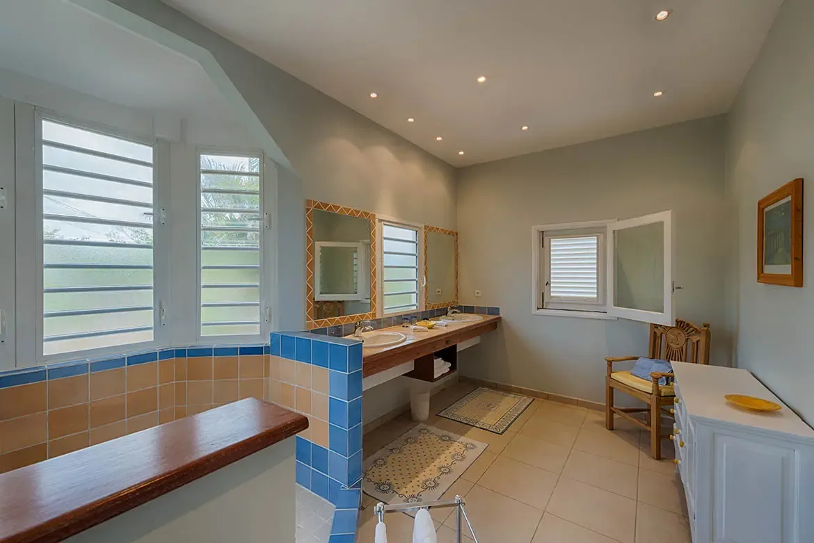 Blue and tan tiles with a zigzag border frame two mirrors above a dark wood double vanity, where a blue-and-terracotta shower partition and a yellow-cushioned chair add Mediterranean warmth.