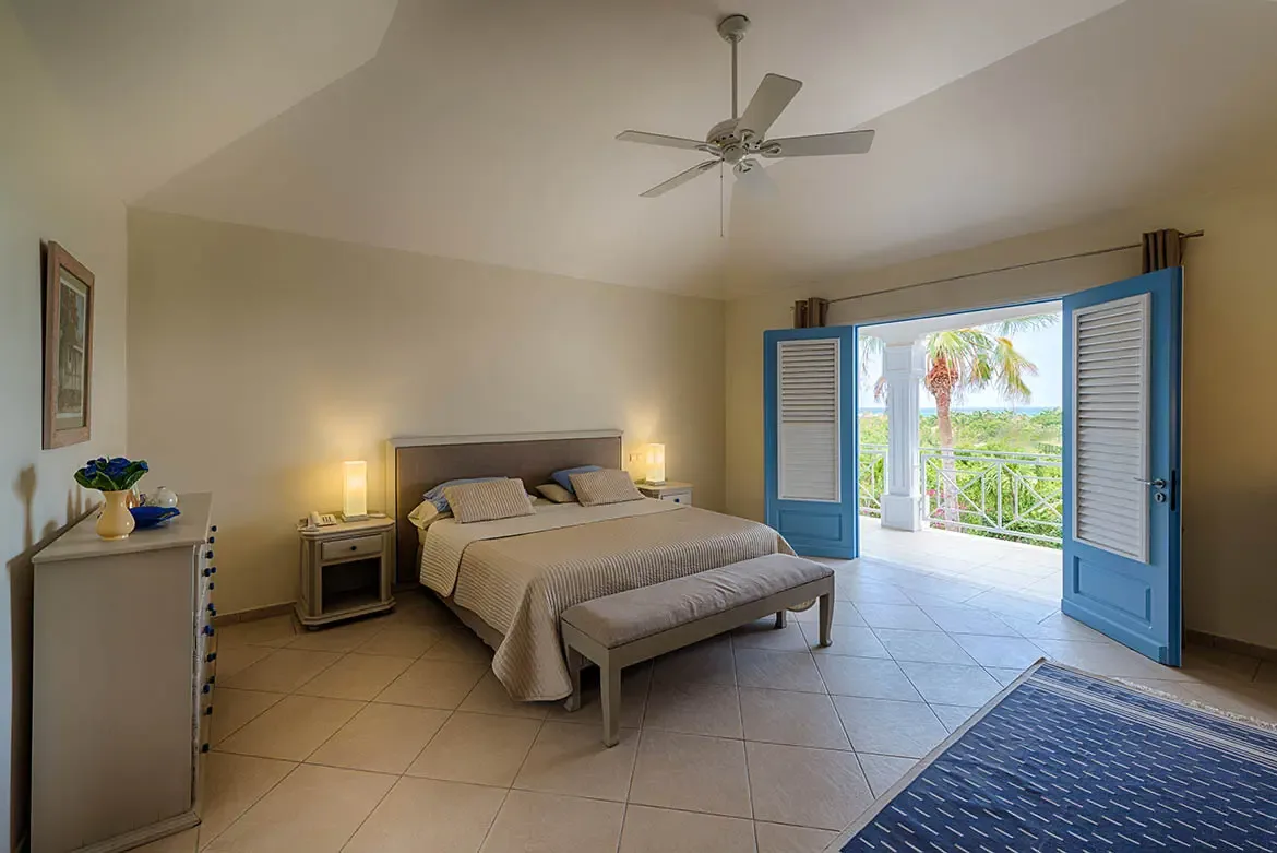 A beige-quilted bed with blue pillows faces bright blue louvered doors that open to a private balcony, where ocean and tropical greenery fill the view beyond white railings.