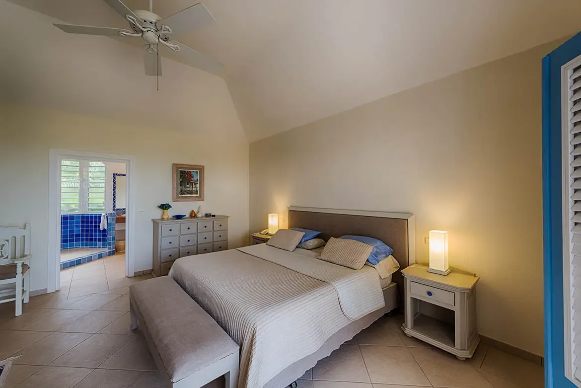 A beige-quilted bed with blue accent pillows sits under a vaulted ceiling, where a white fan spins and an open doorway reveals a blue-tiled bathroom beyond.