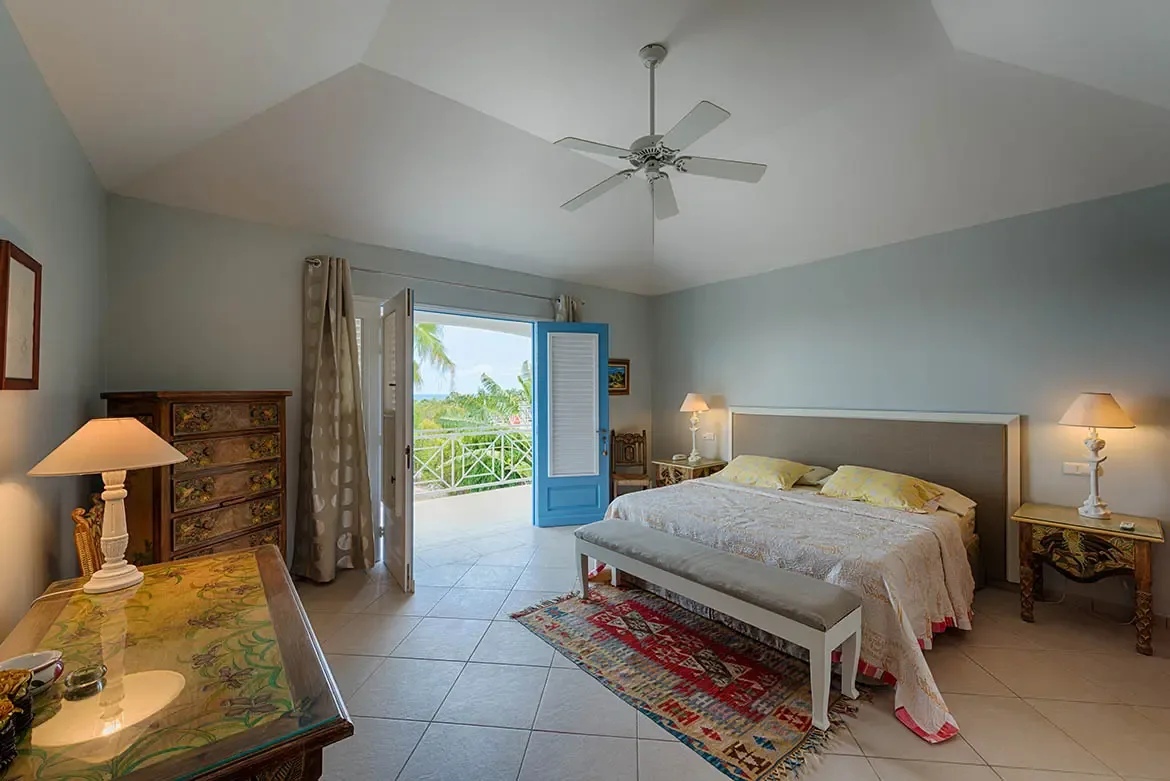 A grey headboard bed with yellow pillows faces blue louvered doors that open to a balcony, where a colorful floral dresser and a painted glass table warm the sunlit room.