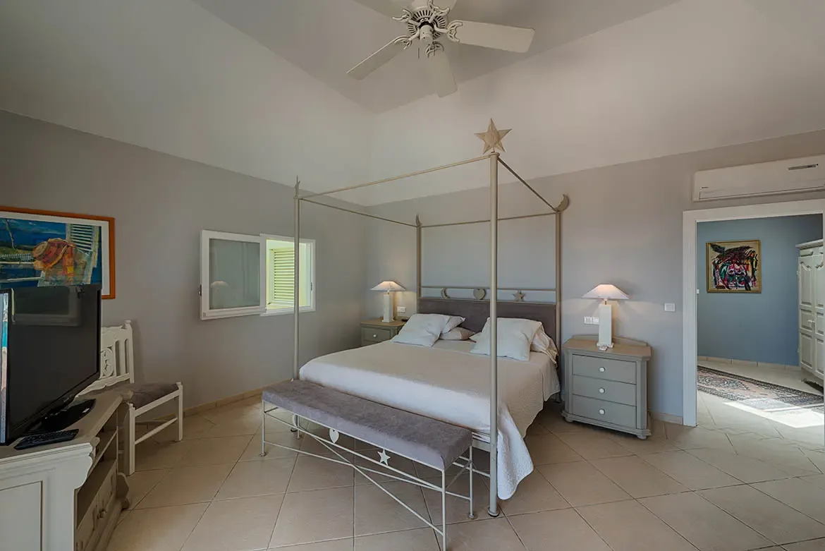 A light-grey canopy bed with a star finial sits on diagonal tiles under a vaulted ceiling, where a rustic TV console and colorful coastal art warm the serene grey-and-white room.