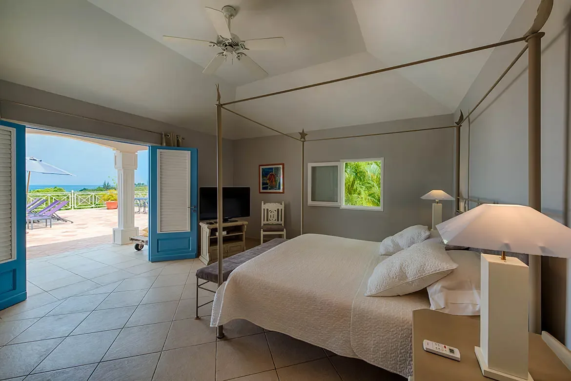 A white-quilted four-poster bed with a metal frame faces bright blue louvered doors that open to a terrace, where purple loungers and an umbrella overlook the ocean horizon.