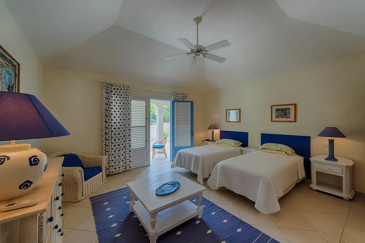 Two twin beds with cobalt blue headboards and green-yellow pillows sit on a navy rug, where a blue-shade lamp and white wicker chair face blue louvered doors to a sunlit patio.