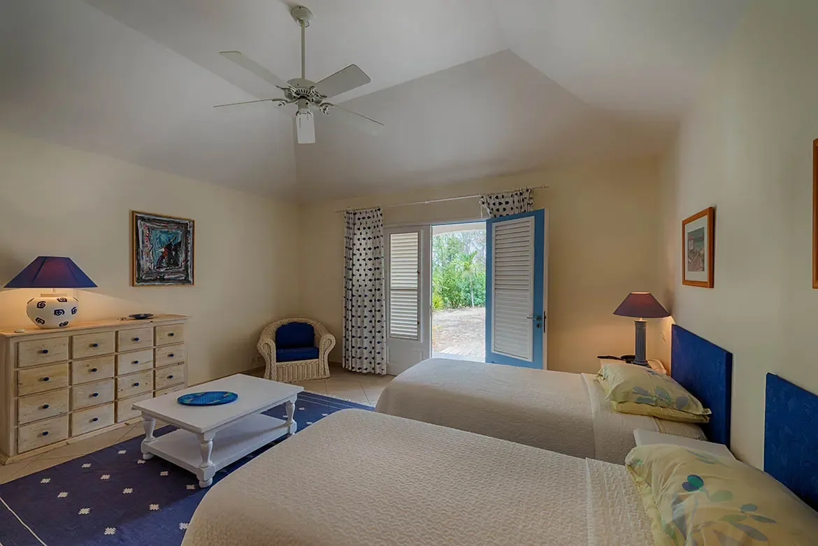 Two white-textured twin beds with royal blue headboards sit on a navy patterned rug, where a light-wood dresser and white wicker chair face blue louvered doors opening to lush greenery.