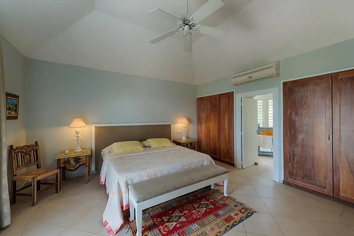 A white bedspread with sunny yellow floral pillows sits on a patterned rug under a vaulted ceiling, where dark-wood built-ins and a white AC unit line the wall near a bathroom doorway.