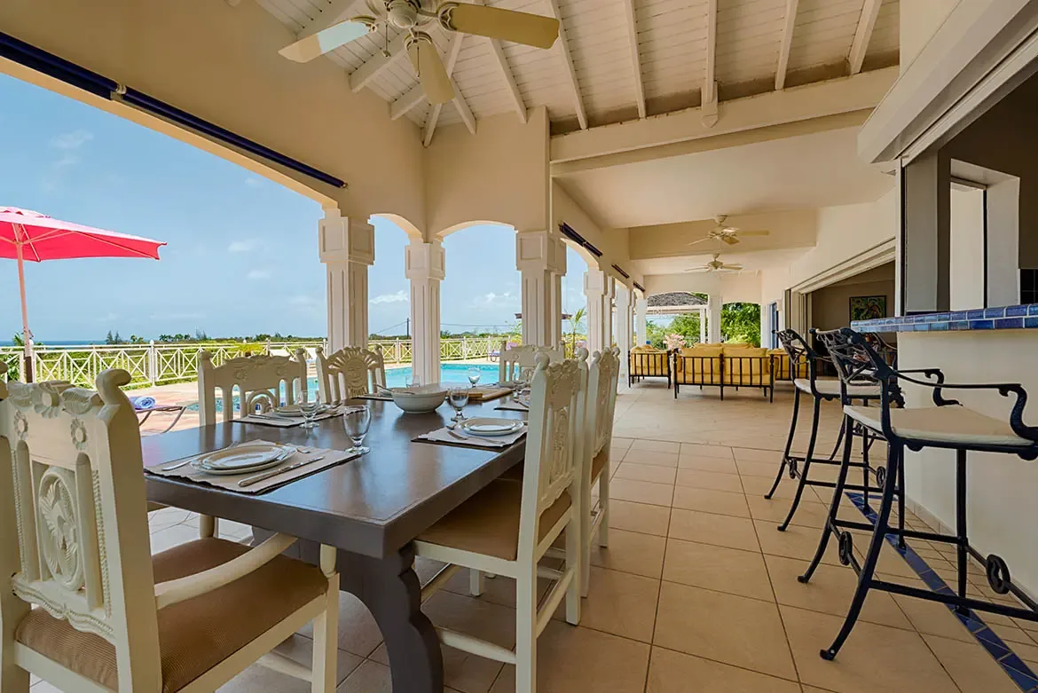 White carved chairs surround a dark wood dining table under a vaulted ceiling, where a blue-tiled bar and yellow sofas open to a pink-umbrellaed pool and ocean view.
