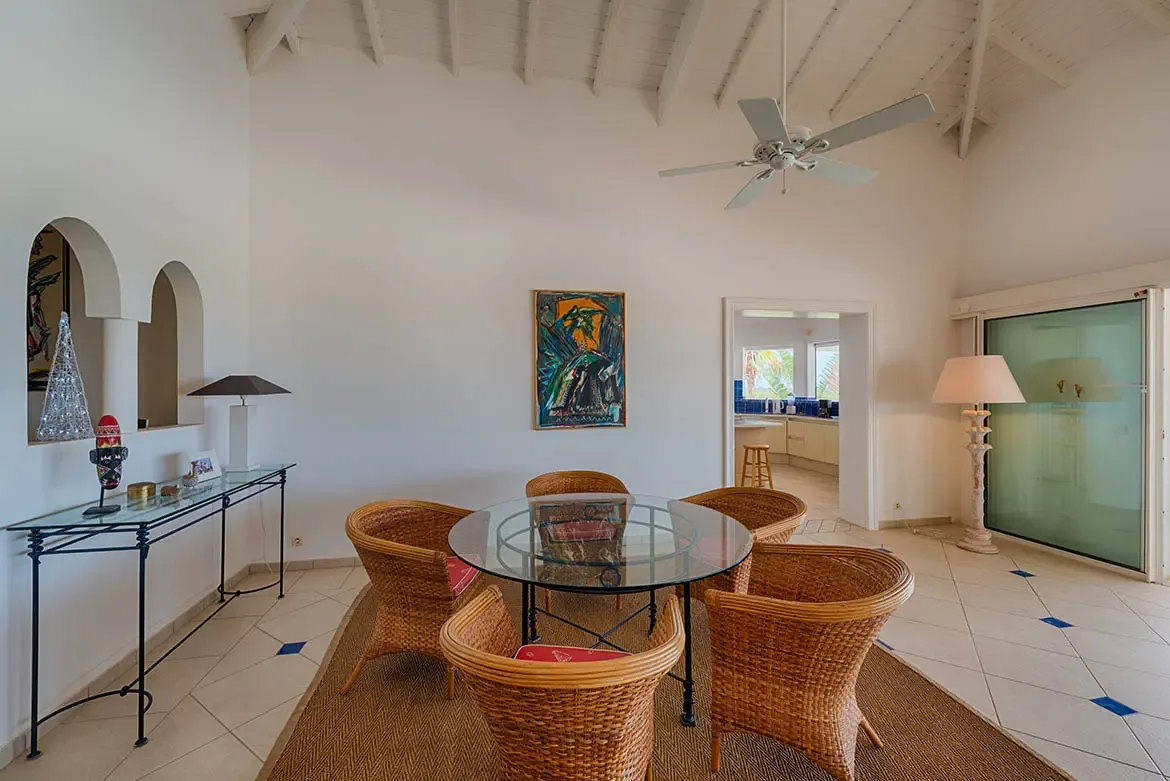 Honey wicker chairs surround a glass table on a sisal rug, where arched niches and abstract art warm the vaulted dining room beside an open kitchen.