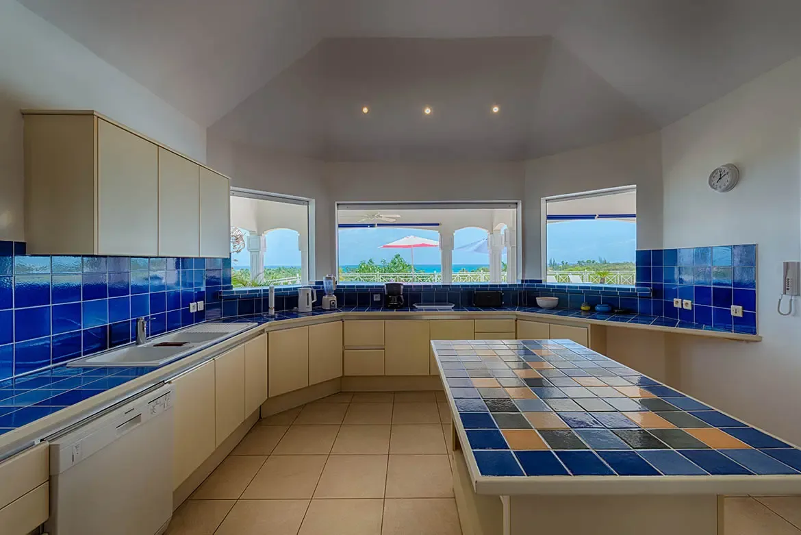 Blue and cream tiles wrap a U-shaped kitchen with a checkerboard island, where three bay windows frame a balcony view of turquoise ocean and lush greenery.