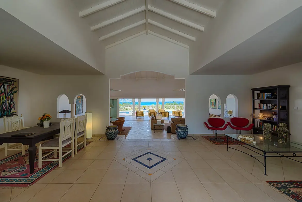 Red pedestal chairs sit on Oriental rugs beside dark bookcases and a glass table, where blue and white ceramic urns flank an archway to a veranda with a turquoise pool and ocean view.