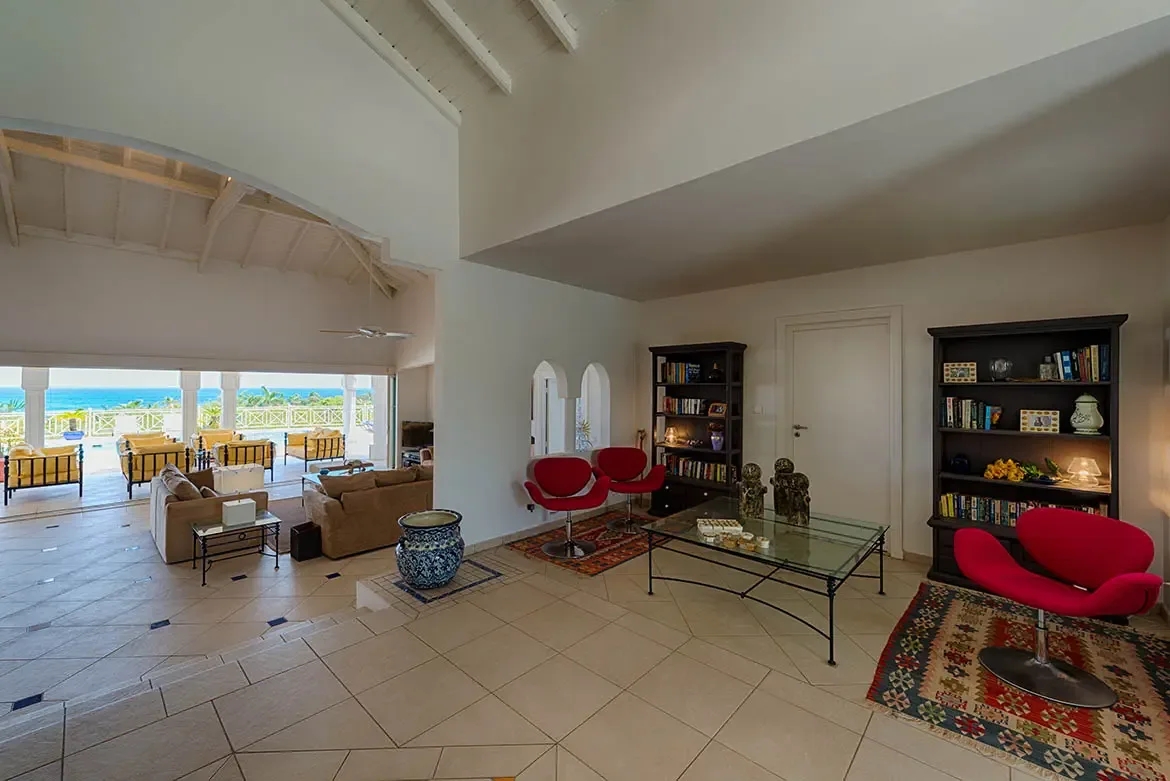 Tan sofas with leopard pillows face a glass coffee table under a vaulted ceiling, where wide doors open to a yellow-cushioned terrace and a turquoise pool above the ocean.