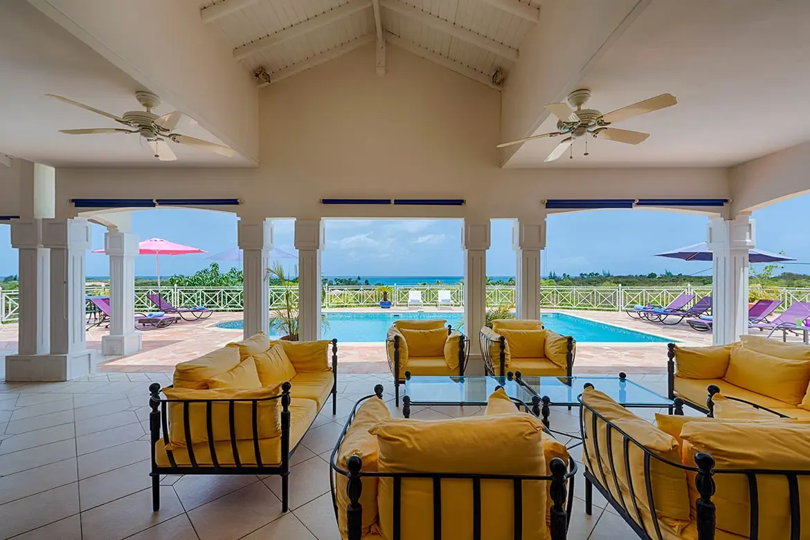 Yellow-cushioned sofas sit under a vaulted ceiling with white columns, where open arches reveal a turquoise pool and ocean beyond lush greenery.