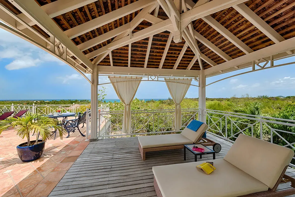 Beige loungers with gauze curtains sit under a white vaulted pavilion, where a blue-potted palm and a turquoise pool overlook lush green hills and ocean.