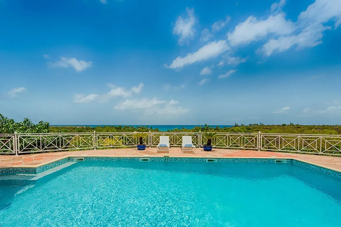 Blue mosaic tiles line a turquoise pool on a terracotta deck, where white loungers between blue ceramic pots overlook lush greenery and a deep blue ocean horizon.