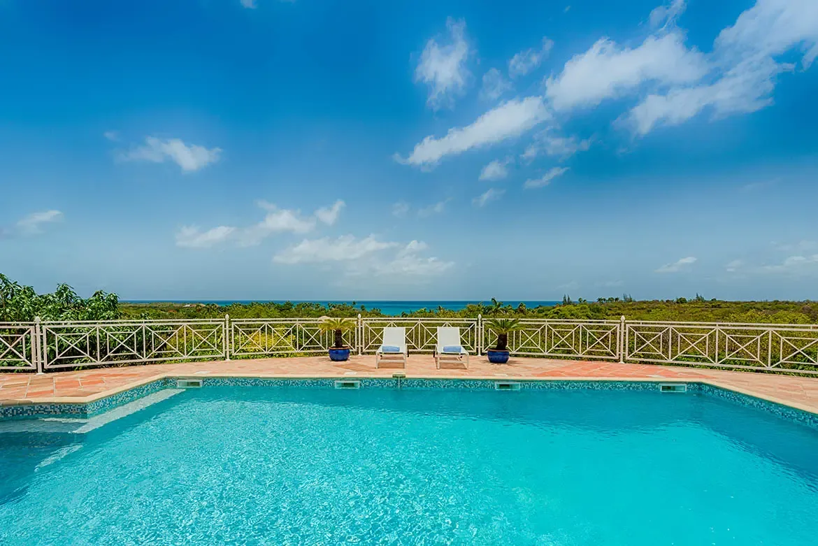 Two white loungers with blue towels sit beside blue ceramic palms on a terracotta deck, where a white railing frames turquoise water and lush tropical hills.