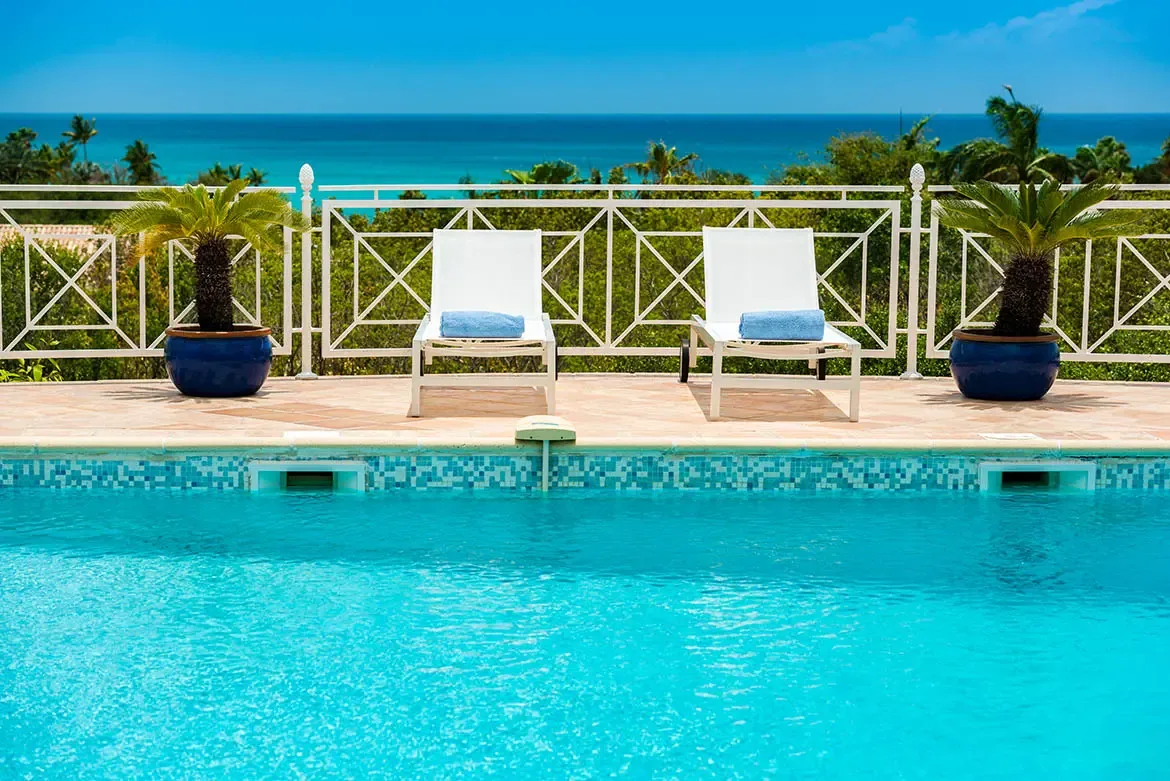 Two white loungers with rolled blue towels sit between glossy blue ceramic palms on a terracotta deck, where a white railing frames a turquoise pool and deep blue ocean beyond.