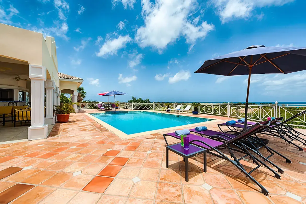 Purple loungers under a blue umbrella sit on terracotta tiles beside a turquoise pool, where yellow sofas shade under white columns and ocean fills the horizon.