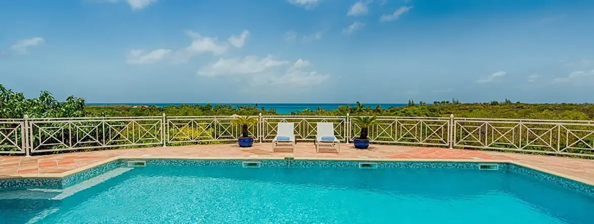 Submerged steps enter a turquoise pool with blue mosaic tiles, where two loungers with blue towels and ceramic palms frame a white railing and ocean beyond.