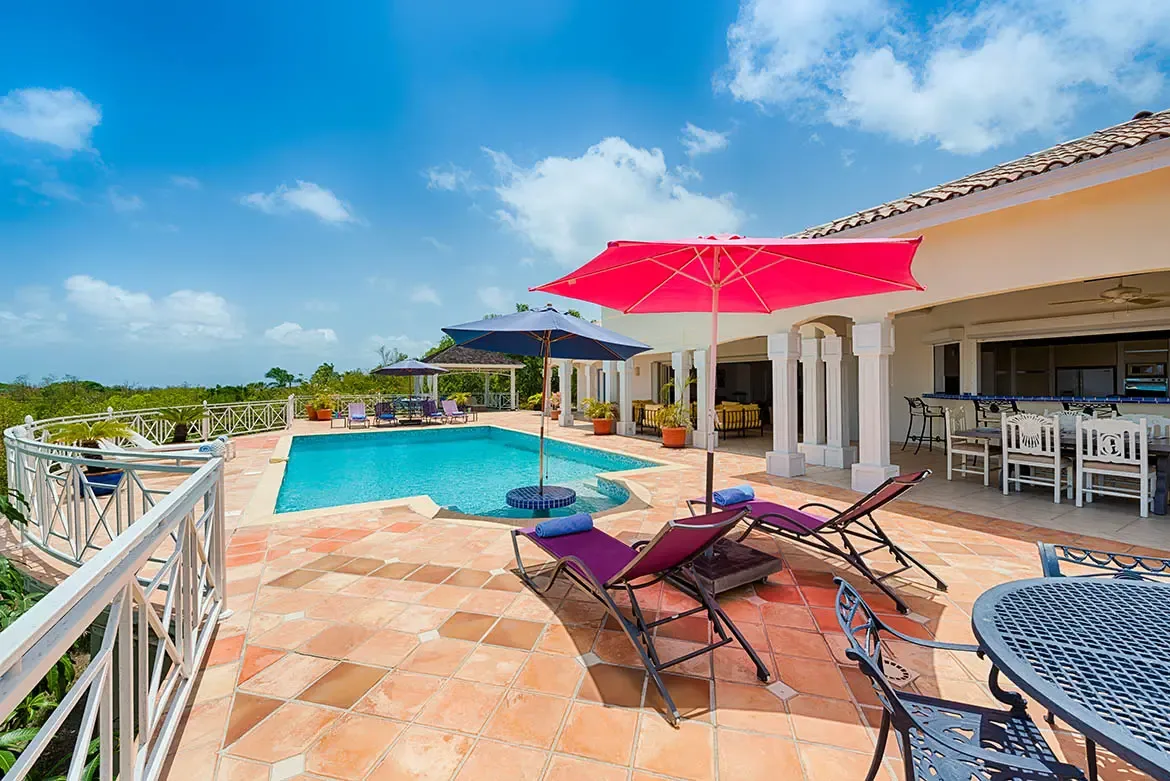 A turquoise pool on a terracotta terrace sits beside a red-roofed villa, where pink and blue umbrellas shade purple loungers and a covered veranda with yellow sofas overlooks the ocean.