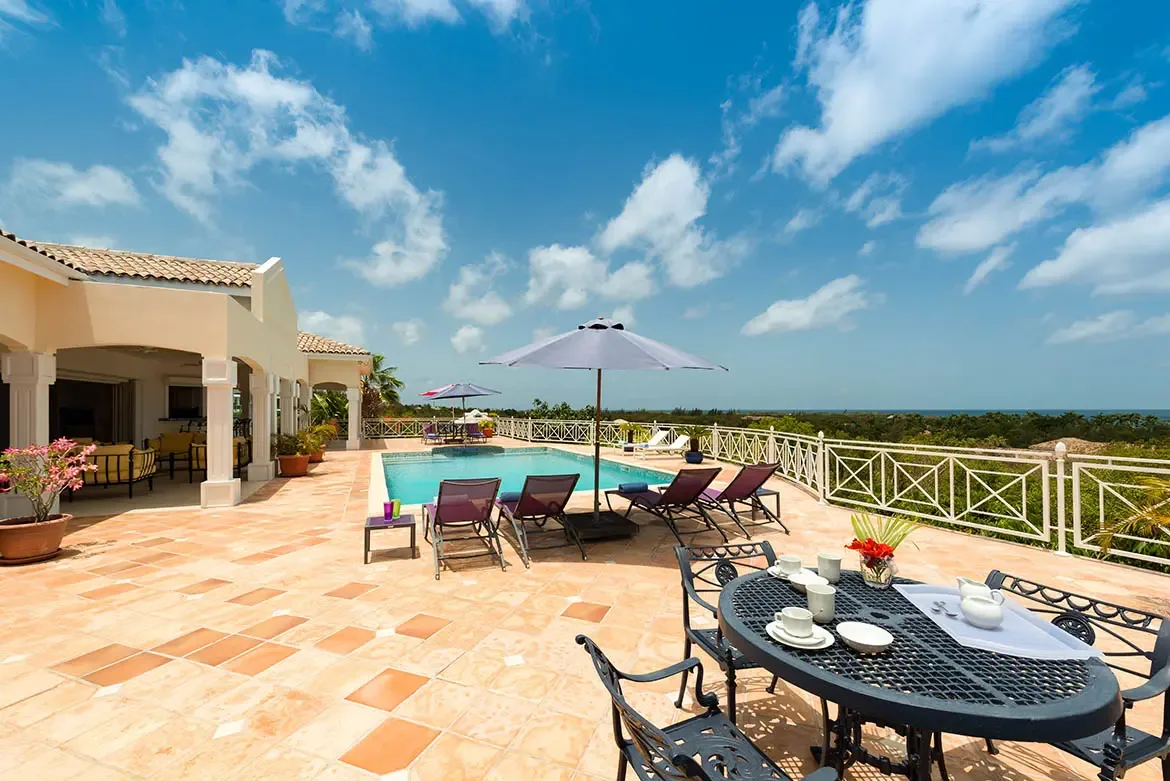 A wrought-iron tea table with red flowers sits on a terracotta terrace, where purple loungers under umbrellas frame a turquoise pool and ocean horizon.