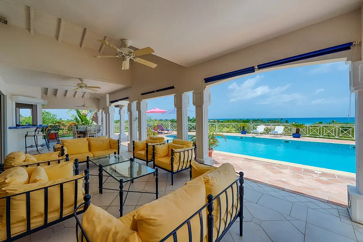 Yellow-cushioned sofas and coffee tables sit under a slatted ceiling with white fans, where arches open to a turquoise pool and ocean view beyond tropical greenery.
