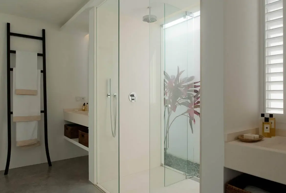 A frameless glass shower features a frosted botanical panel with red-leafed plant motifs and a pebble-lined floor. A black ladder rack holds white towels beside floating sinks with wicker baskets and amber bottles—a spa-inspired bathroom where nature meets minimalism.