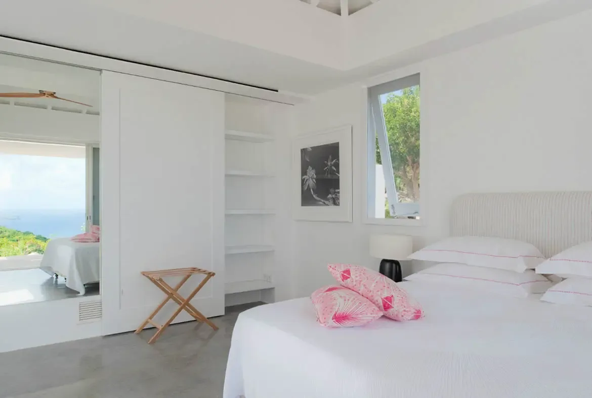 Crisp white linens with pink piped pillows and tropical leaf throw cushions rest on a ribbed headboard. A sliding closet door opens to reveal an ocean and hillside vista beyond—a bedroom where the Caribbean peeks through every corner.