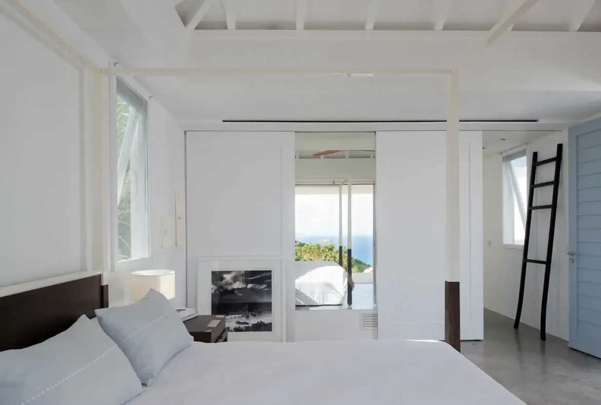 Crisp white linens with light blue pillows sit against a dark brown headboard beneath two black-and-white artworks. White doors open to lush greenery and an ocean glimpse while a leaning black ladder adds industrial edge to this serene coastal bedroom.