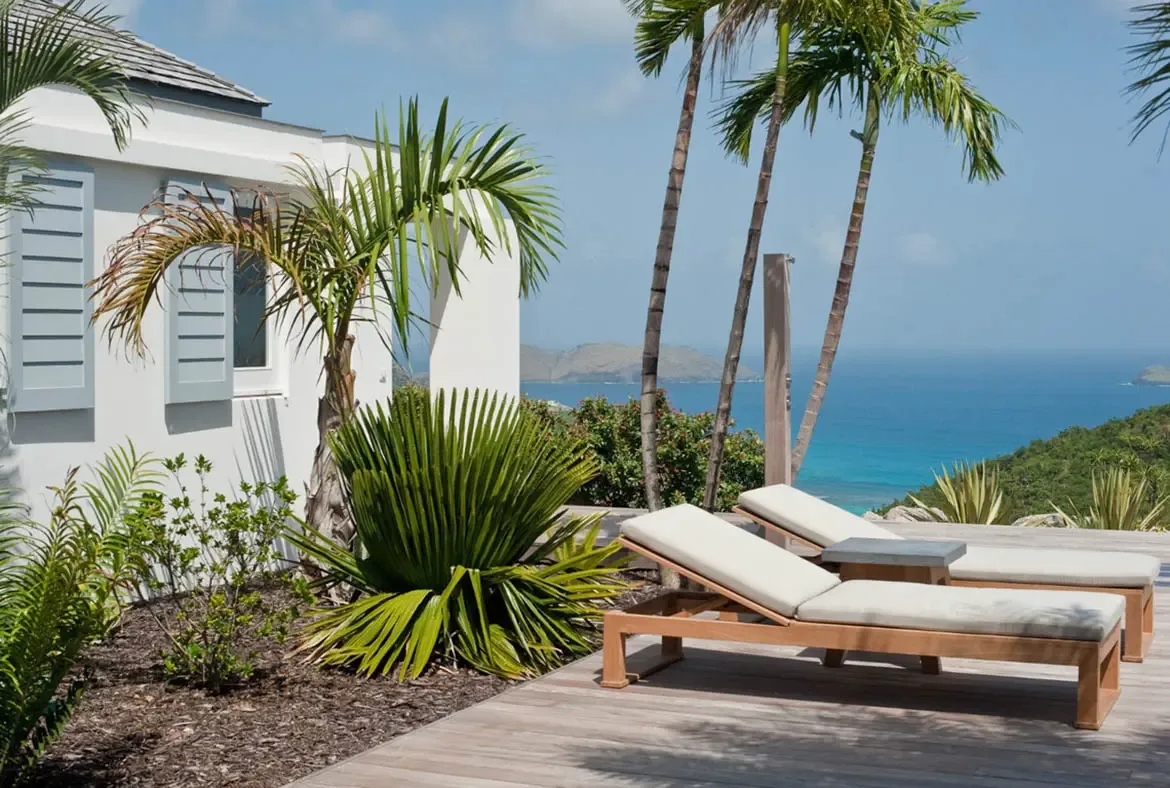 Two plush lounge chairs with beige cushions sit on a warm wooden deck beside a white villa with light blue shutters and a gray shingled roof. Lush palms and tropical foliage frame the turquoise ocean stretching to distant islands under a bright blue sky—pure Caribbean serenity.